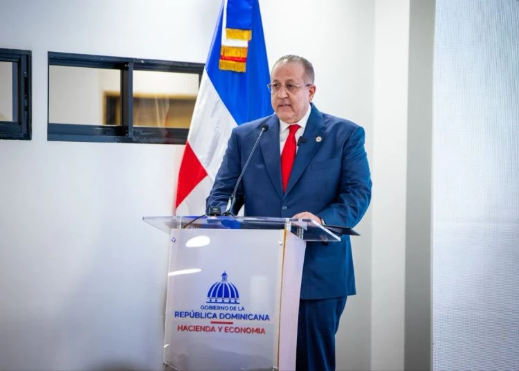 Man in a blue suit and red tie speaks at a clear podium, with the Dominican flag and government seal nearby.
