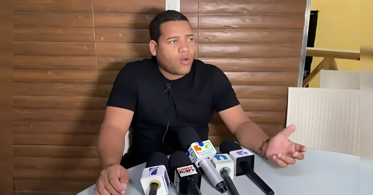 Man in a black t-shirt speaks at a press conference, gesturing with his right hand while microphones sit on the table in front of him.
