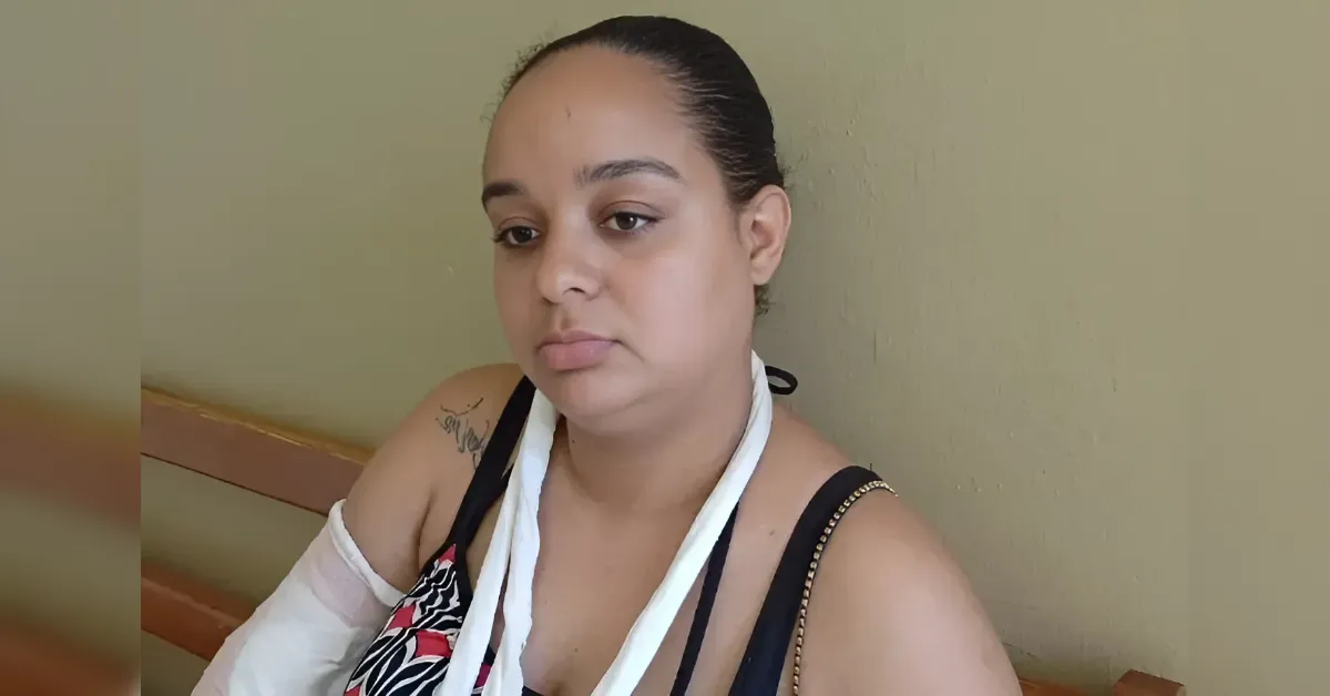 Woman with an injured arm in a white sling resting on her shoulder, seated against a beige wall, looking downward.