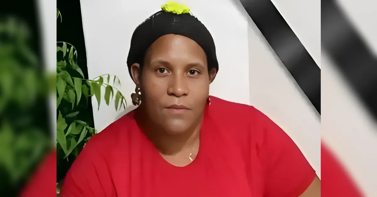 Woman with a black head wrap and red shirt, looking at the camera in front of a white wall with a black ribbon.