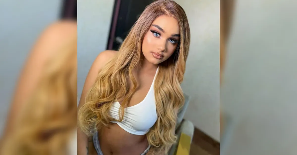 Young woman with long wavy blonde hair in a white crop top, looking at the camera indoors.