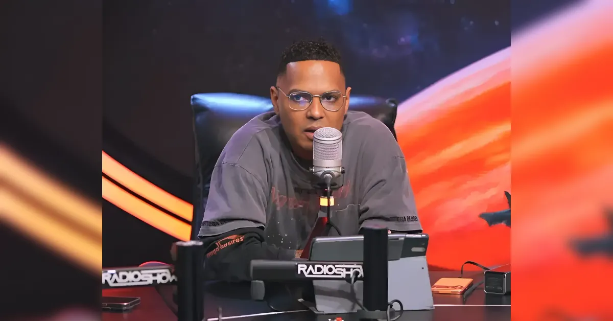 Man wearing glasses speaks into a microphone on a radio show set, with brand signage visible on the desk.