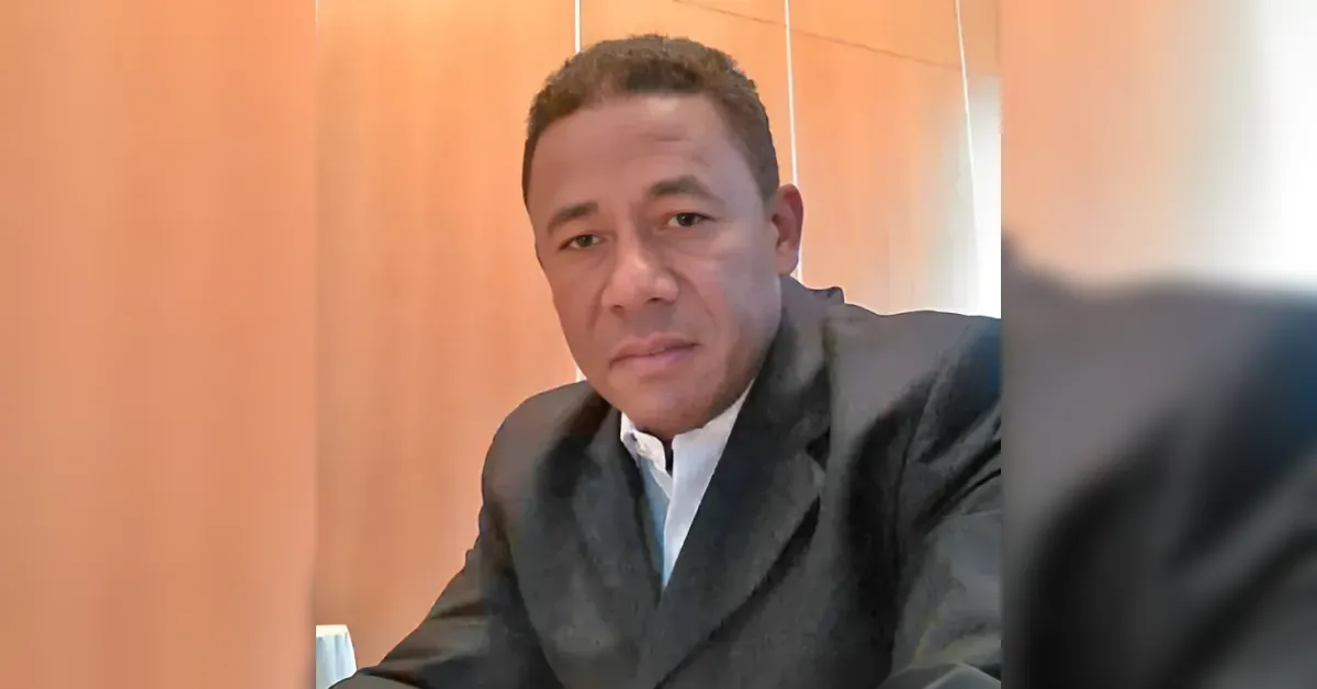 Man in a dark blazer and white shirt looks at the camera in a wood-paneled room, possibly during a video call.
