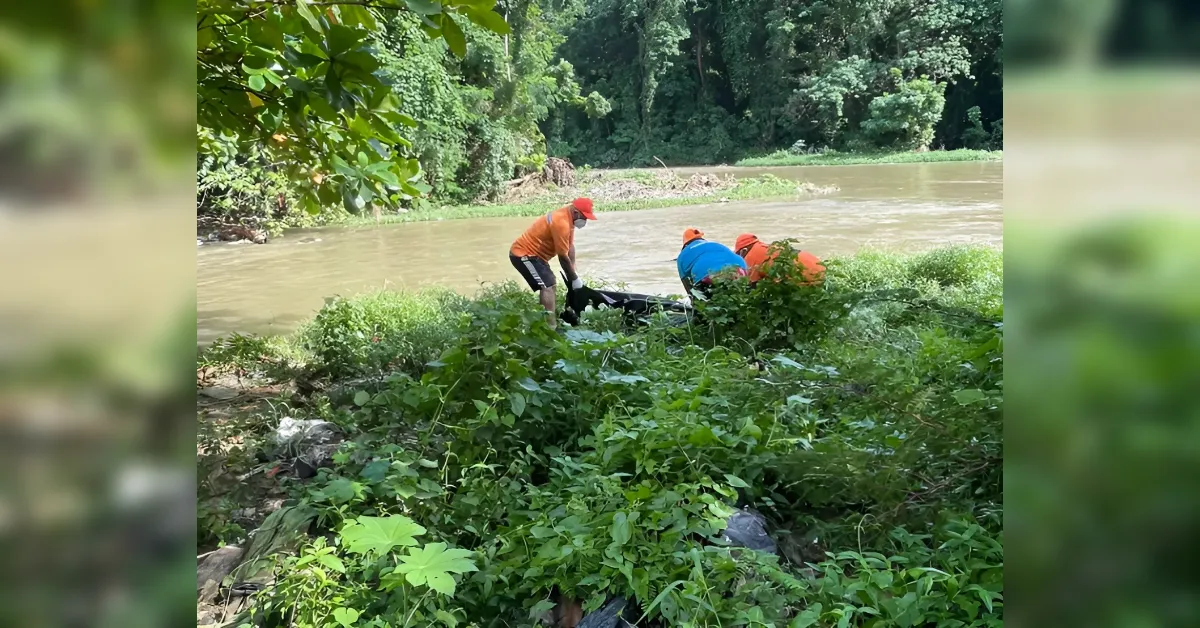 People wearing orange and blue shirts cleaning a muddy riverbank, collecting trash into bags amid green vegetation.