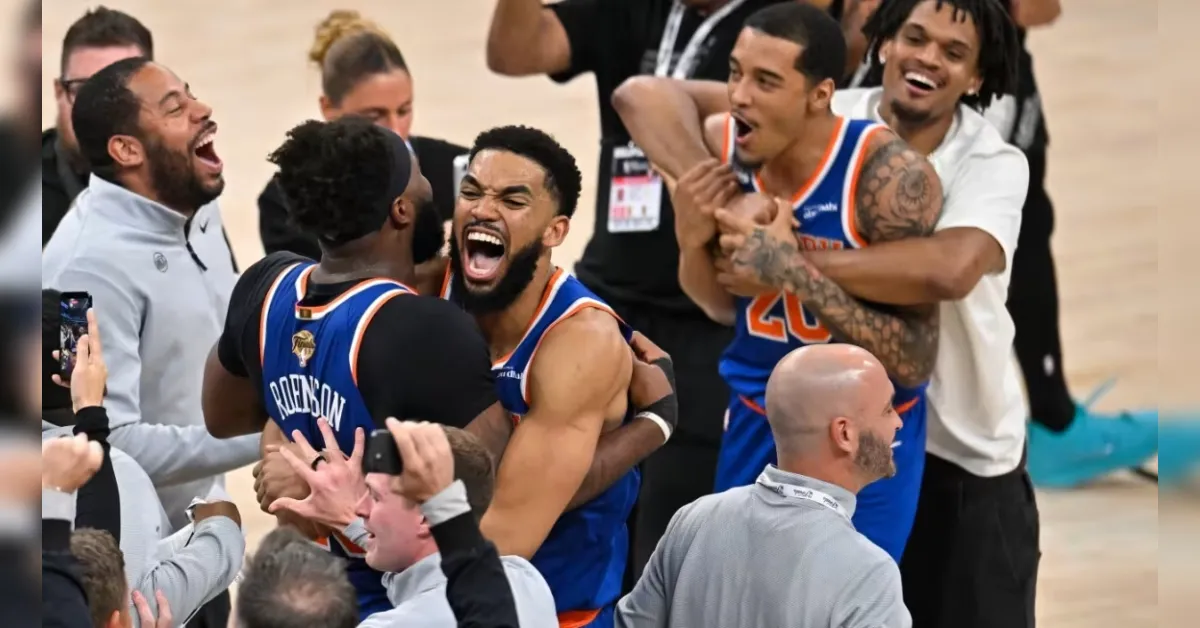 Basketball players in blue and orange jerseys celebrate together, embracing and cheering amid fans and photographers in a gym.