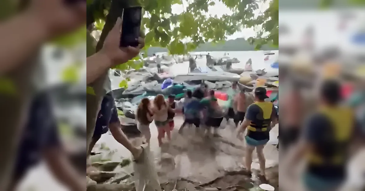 Group of people wearing life jackets gather on a rocky shoreline beside damaged boats and a collapsed dock, while a person on the left holds a phone up to record.