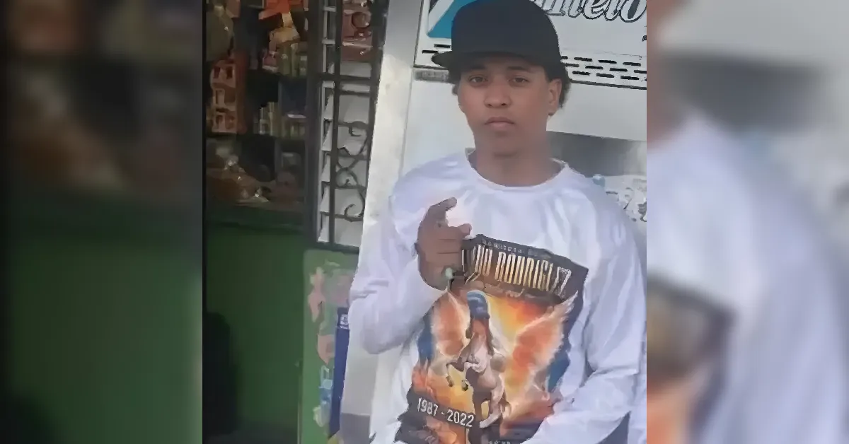 Young man wearing a black cap and white long-sleeve shirt points at the camera while holding a colorful poster outside a shop.