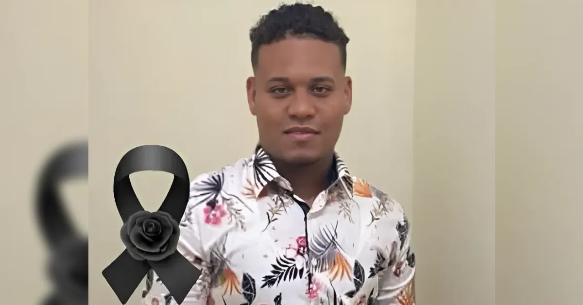 Portrait of a young man in a white floral shirt standing against a plain wall with a black mourning ribbon emblem on the left, signaling a tribute.