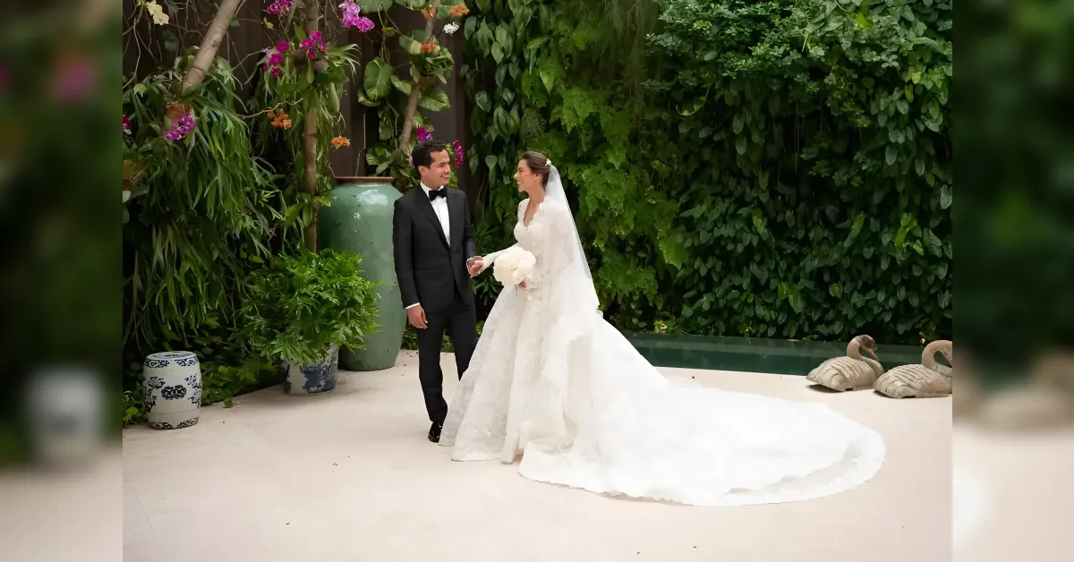 Bride and groom in formal wedding attire walking in a lush garden setting, the bride holding a bouquet and veil trailing behind her.