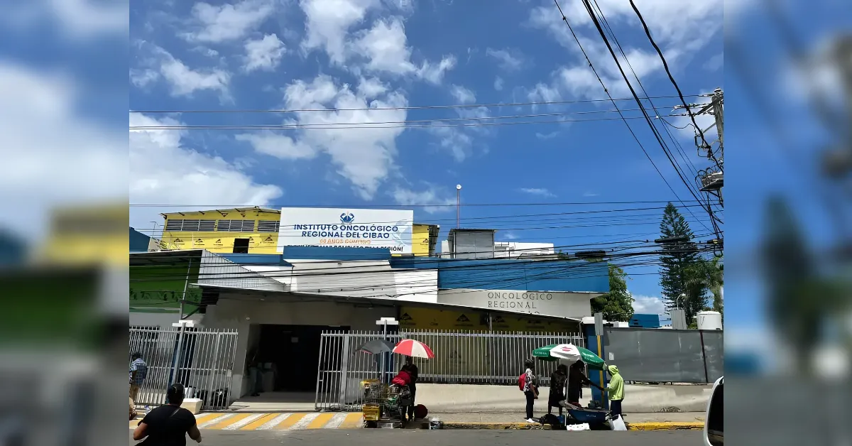 Front view of a regional oncology institute with a blue sign reading 'INSTITUTO ONCOLOGICO REGIONAL DEL CIBAO', metal gates, and power lines overhead.