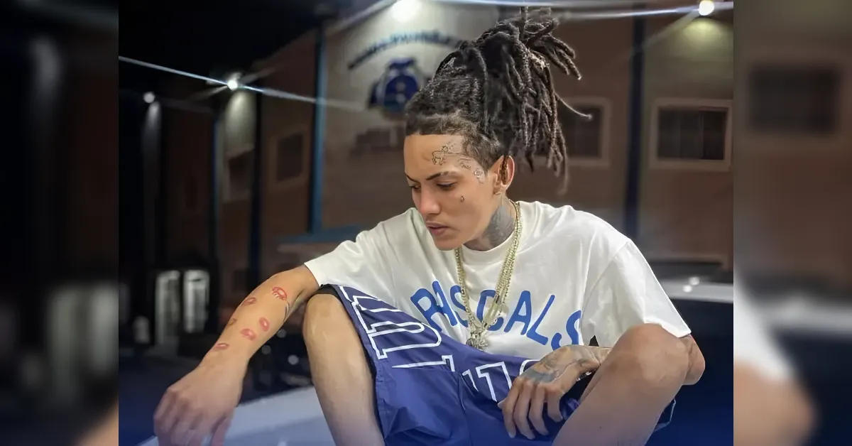 Portrait of a young man with long braided hair, tattoos on his face and arms, wearing a white T-shirt and blue shorts, seated with a leg up in a dim indoor space, looking downward.