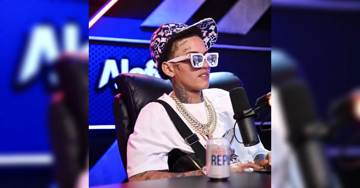 Person wearing a floral cap, oversized white sunglasses, and chunky gold chains sits at a podcast mic in a studio, with a can on the table in front of them.
