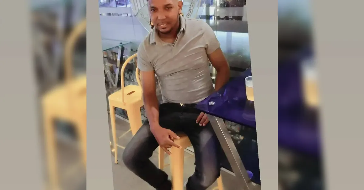 Man seated at a cafe counter on a yellow stool, wearing a light gray polo and dark jeans, with a purple napkin on the table behind him.