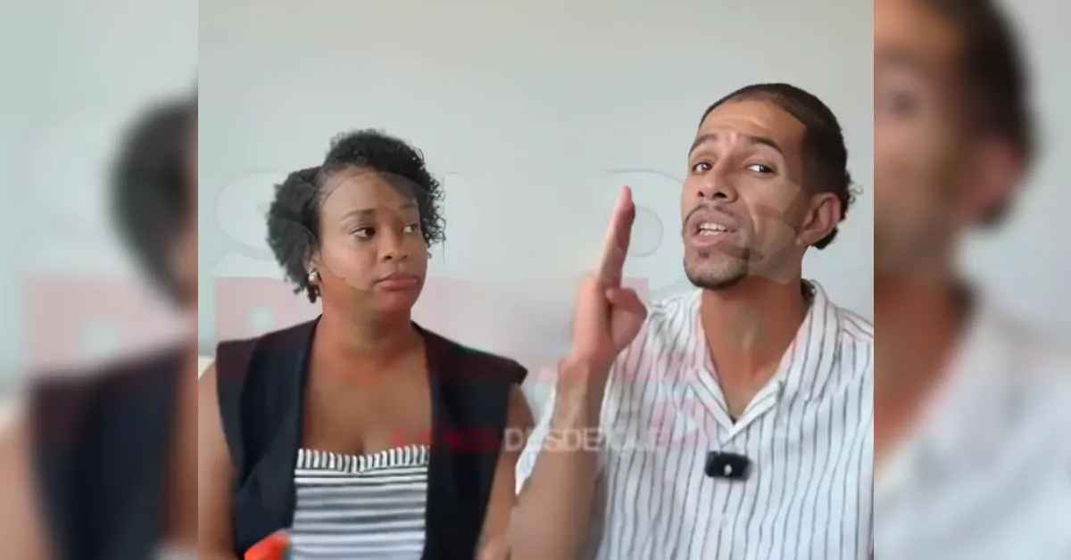 Two people sit indoors with facial masks on; the man speaks animatedly with a raised finger while the woman looks on skeptically.