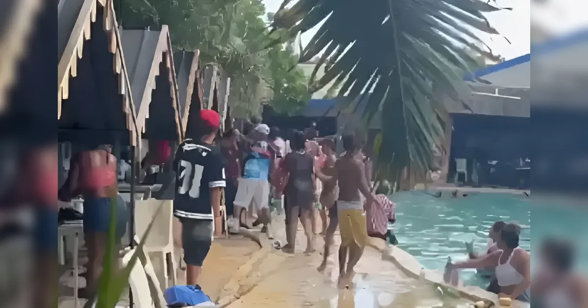 Crowd of people walking along a poolside deck at a tropical resort with thatched cabanas and palm trees nearby.