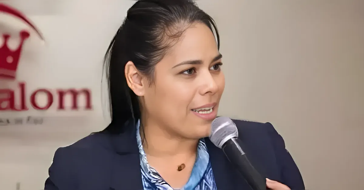 Woman in a navy blazer speaks into a handheld microphone during a panel discussion, with a blurred logo in the background.