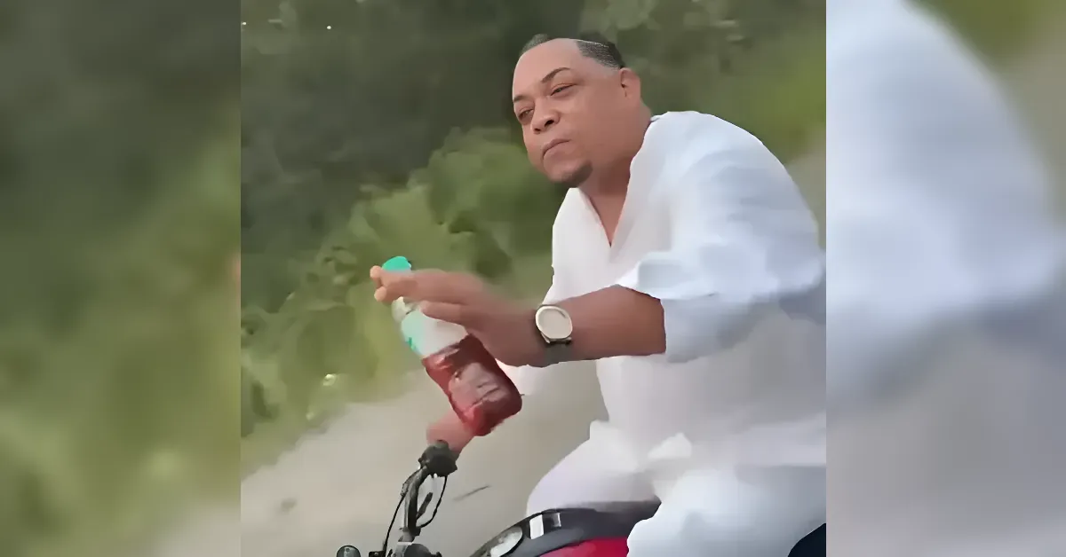 Man in a white shirt pours a red beverage from a plastic bottle while riding a motorcycle on a sunny, rural road.