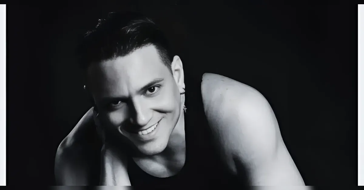 Black-and-white portrait of a smiling man in a sleeveless shirt leaning toward the camera against a dark background, studio lighting.