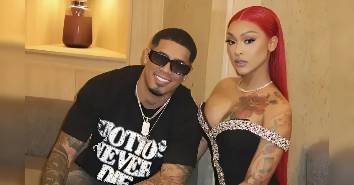Smiling man in a black tee and sunglasses sits beside a woman with bright red hair in a rhinestone-detailed black dress, tattoos visible in the background.