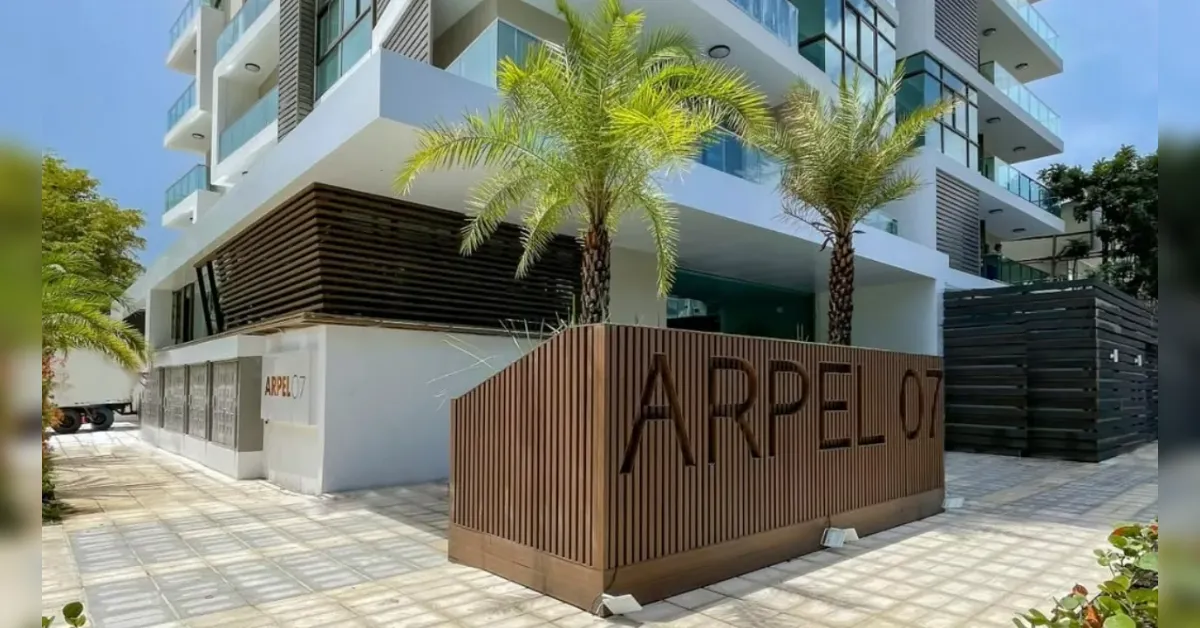 Modern white apartment building with glass balconies and palm trees; a brown slat fence reads ARPEL 07 in the foreground.