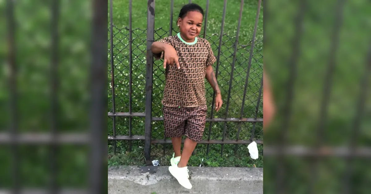 Child posing by a metal fence, wearing a brown matching patterned outfit and bright white sneakers.