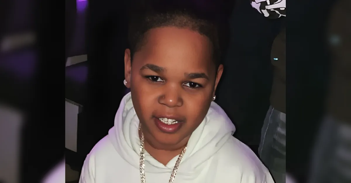 Young person with short hair wearing a white hoodie and silver chain, looking at the camera in a dimly lit setting.