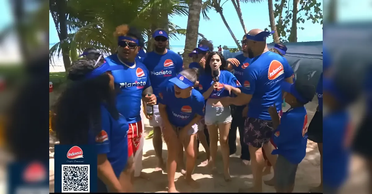 Group of people in blue shirts with Planeta logos on a sunny beach, one person holding a microphone for an interview/announcement.