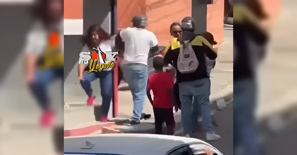 Group of people on a sidewalk gathered around a person with a helmet and backpack, with a child in a red shirt nearby.
