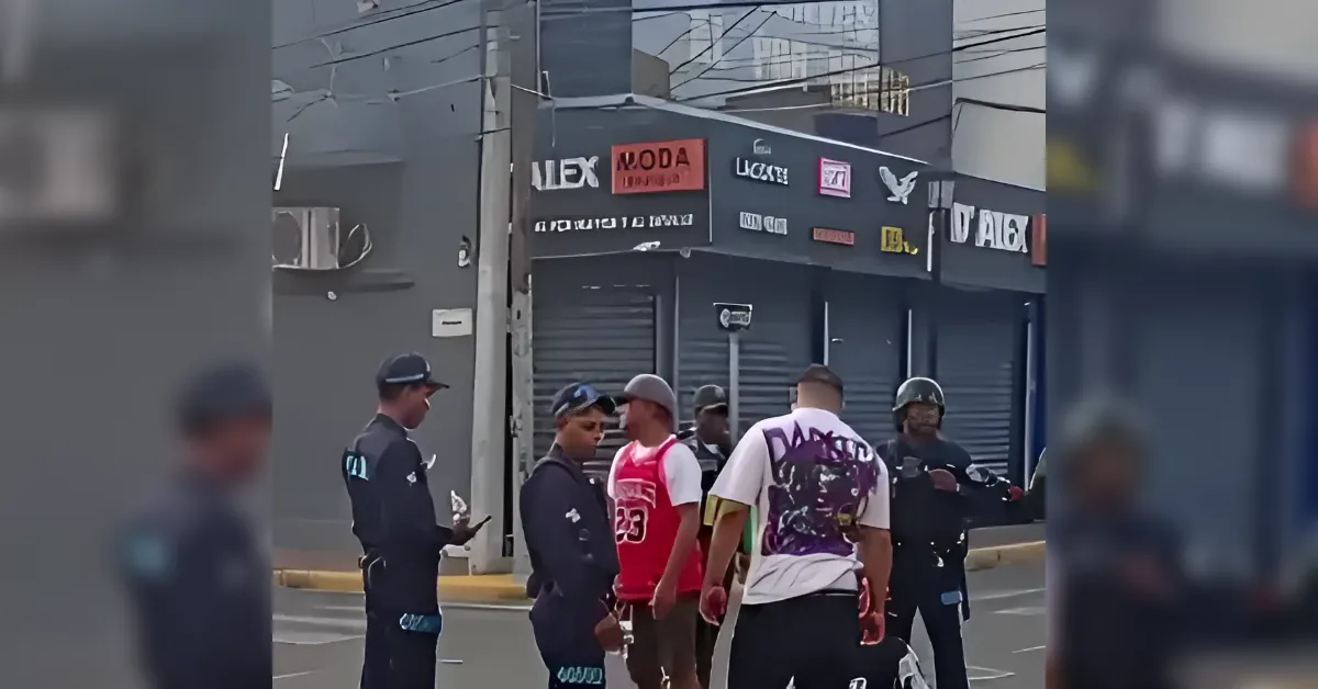 A group of duty police or security officers standing in the street and talking to a few men near a gray storefront with closed metal shutters and various signs on the facade; daytime urban scene.