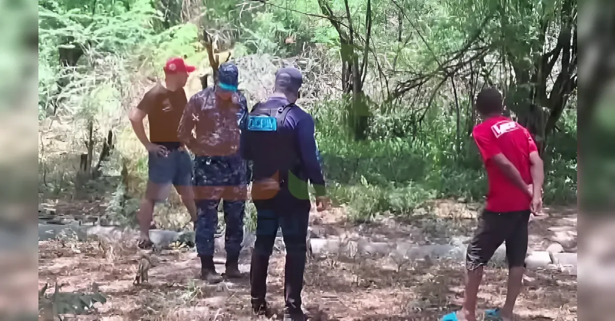 Group of men in a wooded clearing; a uniformed officer talks to two men, while a third man in a red shirt stands with his hands behind his back.