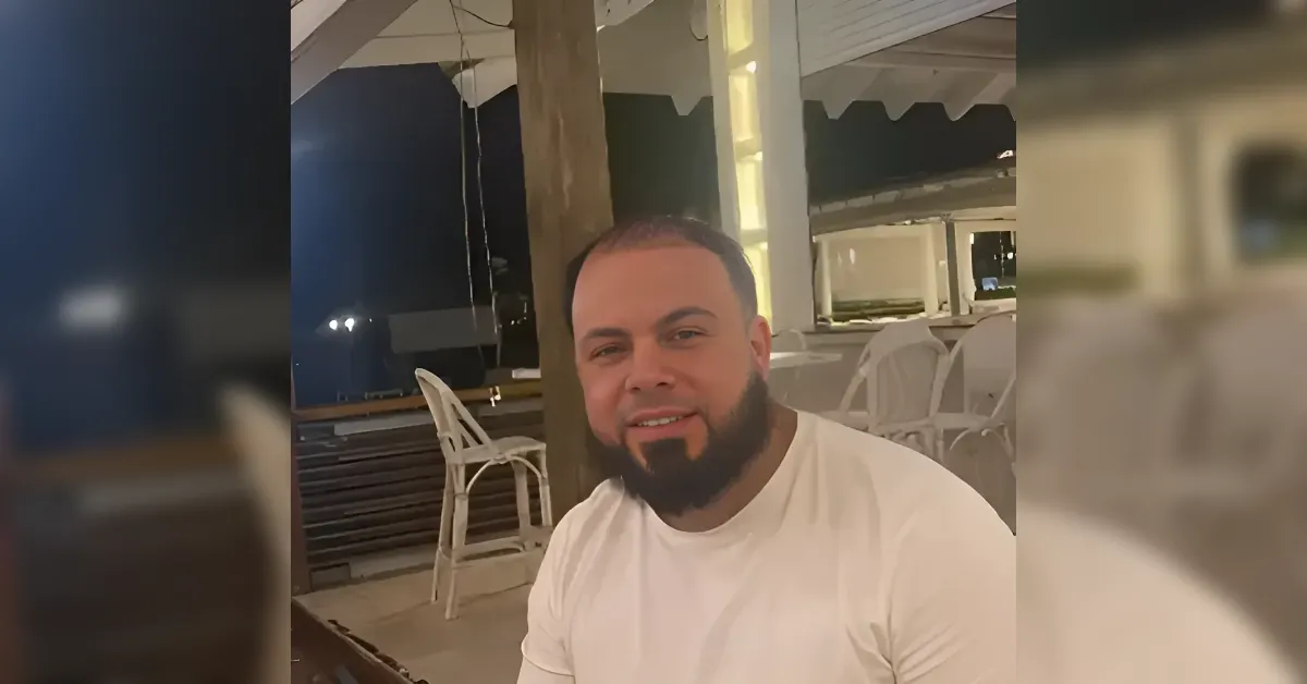 Man with short hair and a full beard sits at an outdoor restaurant patio at night, wearing a white t‑shirt with white chairs in the background.