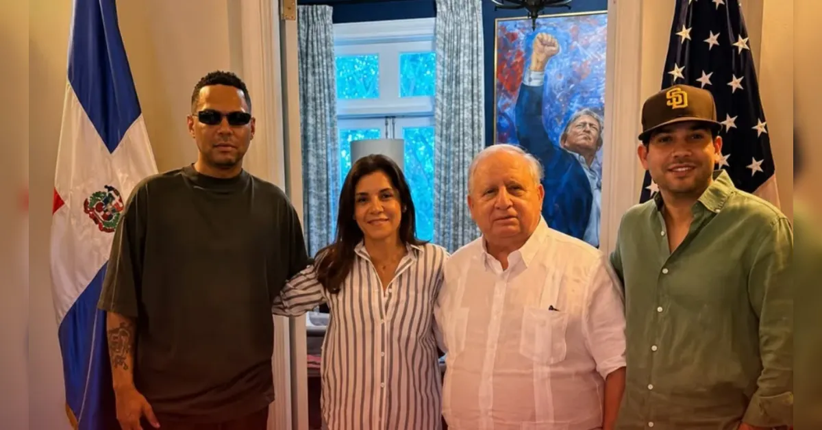 Four people pose for a photo indoors, with the Dominican flag on the left and the U.S. flag on the right behind them.