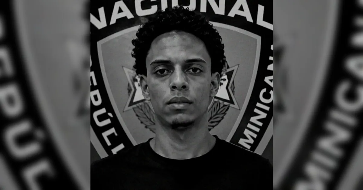 Mugshot of a young man with curly hair, wearing a black shirt, standing in front of a police badge backdrop (black-and-white).
