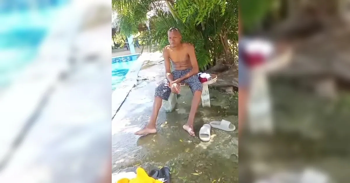 Shirtless man in patterned shorts sits on a small bench by a pool, barefoot with sandals on the wet ground.