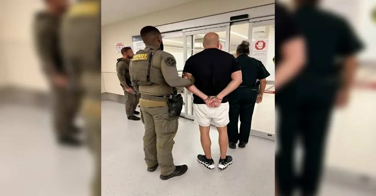 Handcuffed man in black shirt and white shorts is escorted by two officers in a hallway.