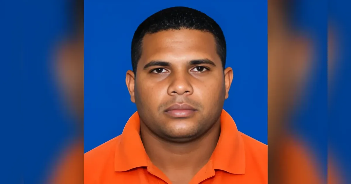 Portrait of a man with short black hair wearing an orange polo shirt against a blue background