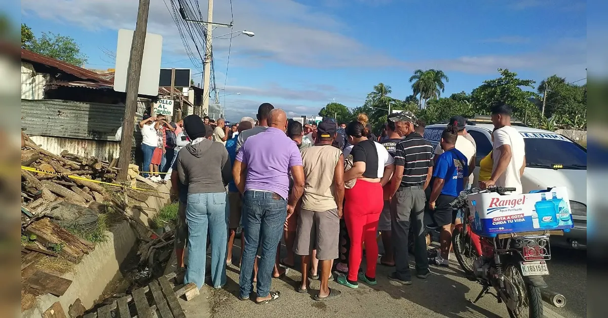 Group of people standing on a street near a pile of broken wood and a taped-off area, watching something off-camera.