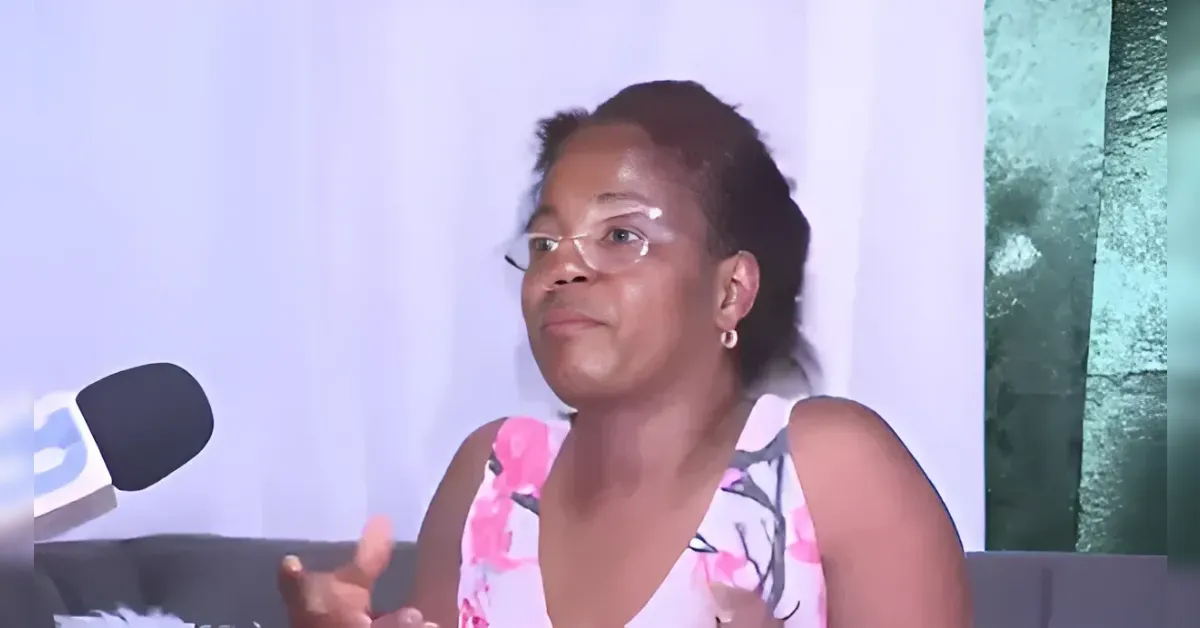 Woman in a pink floral sleeveless top speaks during an interview, gesturing with her hands as a microphone sits to the left.