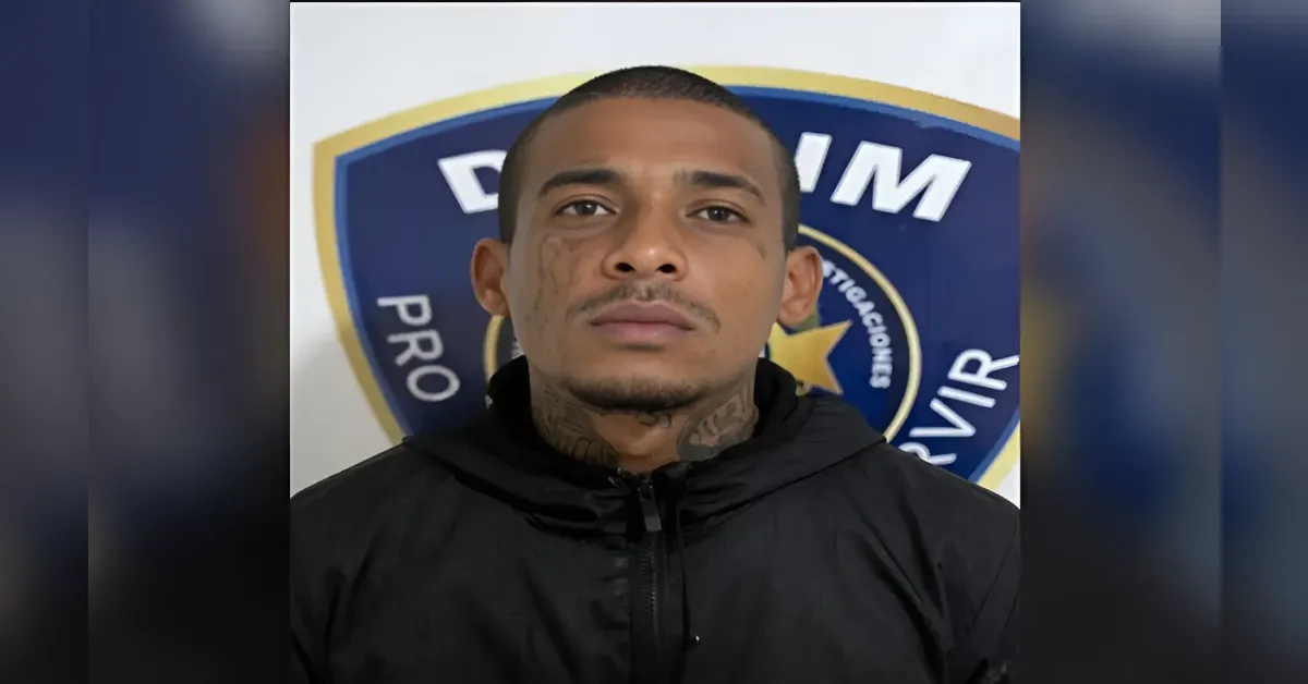 Mugshot-style photo of a man with neck tattoos wearing a black jacket, standing in front of a blue and gold police-style badge backdrop