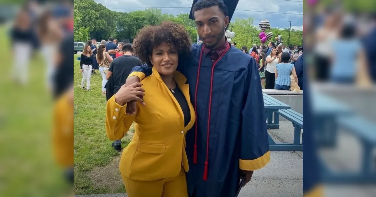 Graduate in a blue gown and cap posing with a person in a bright yellow suit at an outdoor graduation ceremony, with a crowd in the background.