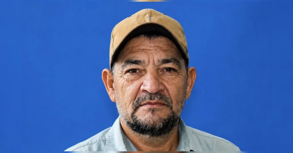 Middle-aged man wearing a beige cap and light blue shirt against a solid blue background, with a neutral expression.