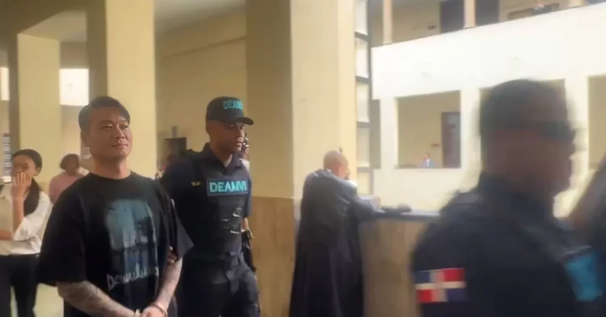 Man in a black graphic t-shirt with tattoos stands with hands cuffed, escorted by a security officer in a dark uniform through a public building corridor.