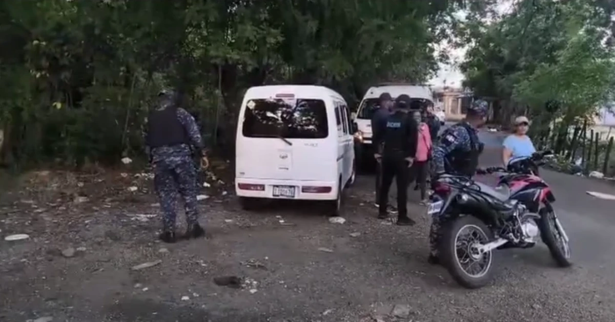 Group of people near a white van on a gravel road, with a motorbike parked nearby and others standing around under trees.