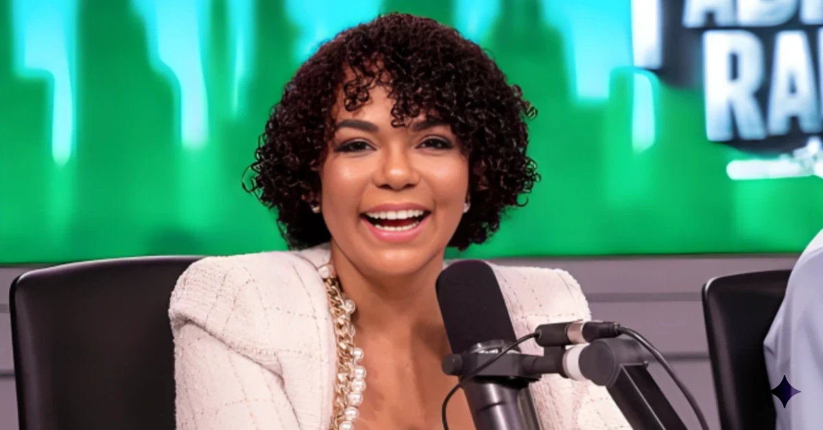 Woman with curly hair smiles while speaking into a microphone in a studio with a green backdrop.