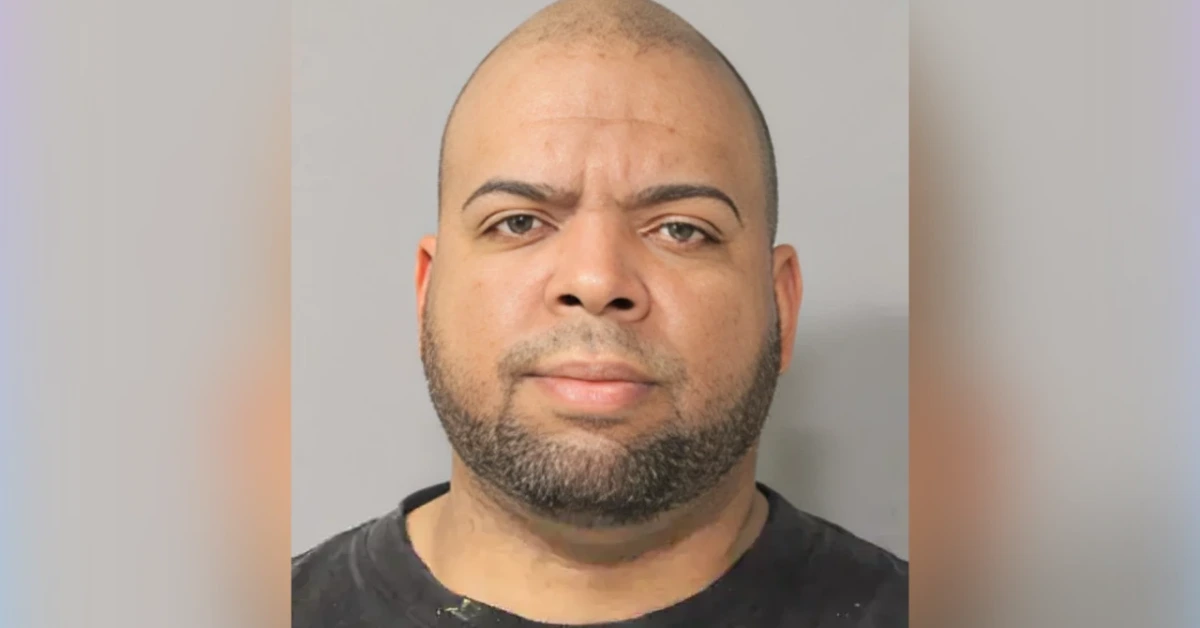 Front-facing mugshot-style portrait of a man with a trimmed beard and shaved head against a gray background.
