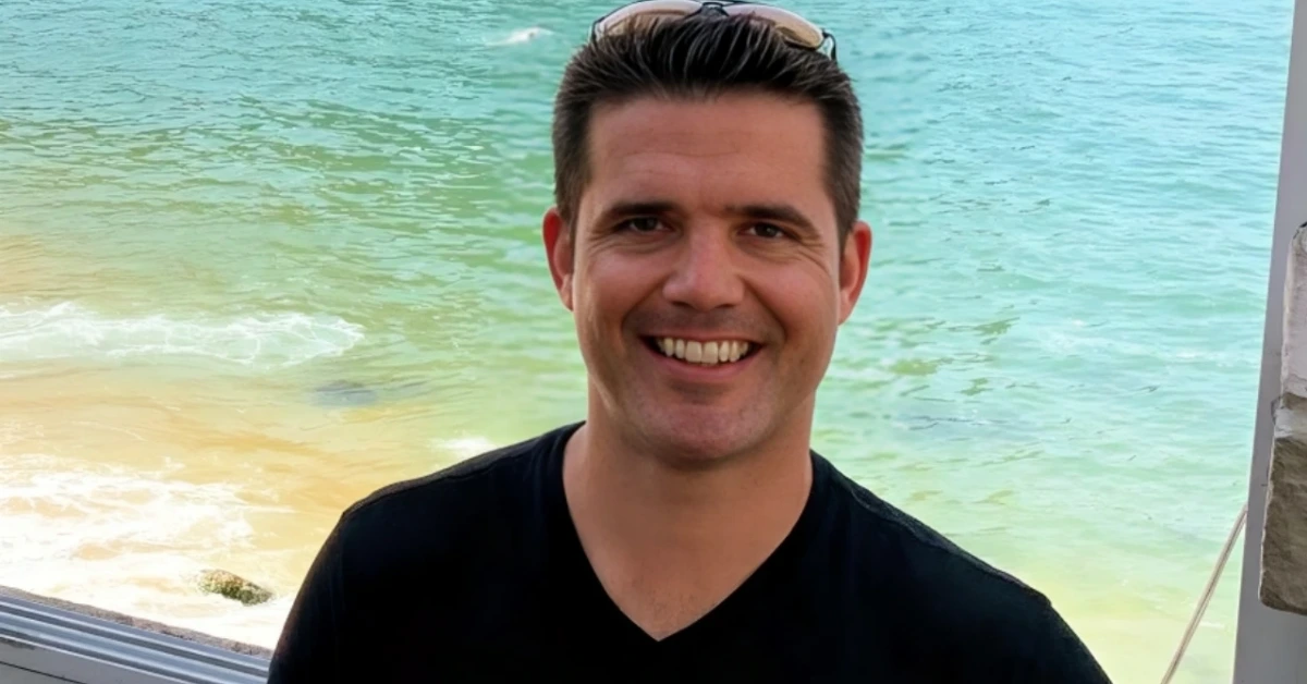 Smiling man in a black V-neck shirt with sunglasses on his head, standing by turquoise ocean water and a sandy beach behind him.