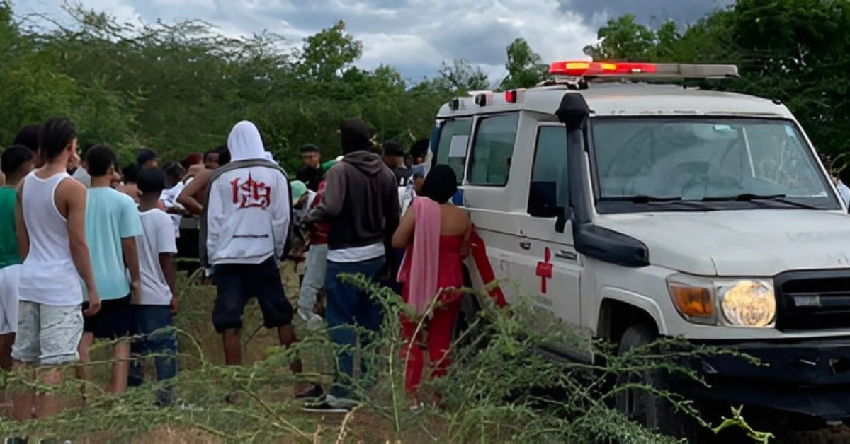 Ambulance with red cross and flashing lights as a group of people gathers nearby in an open rural area.