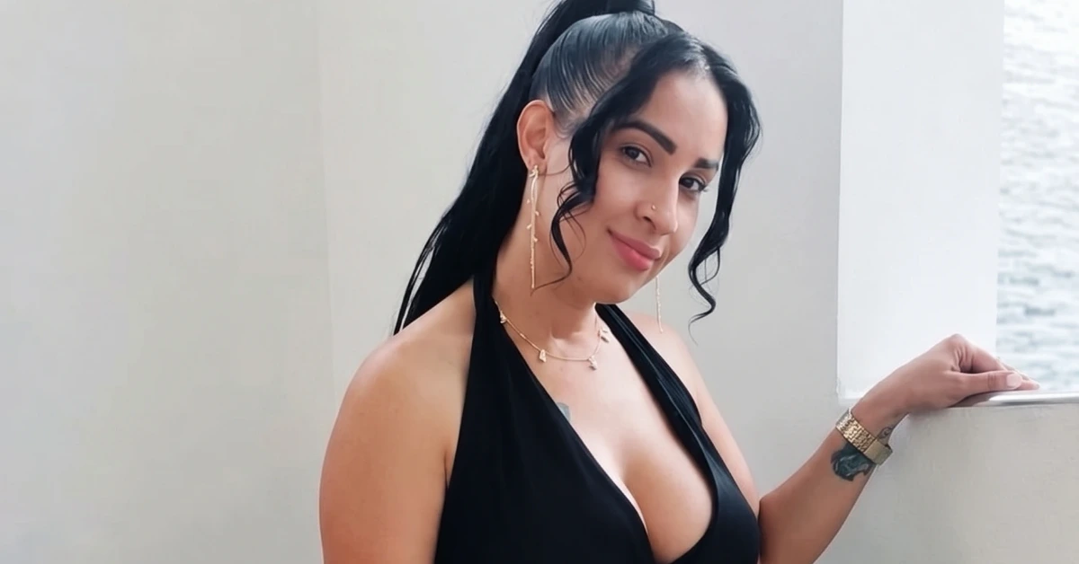 Smiling woman with long black hair in a black halter top, leaning on a railing beside a body of water.