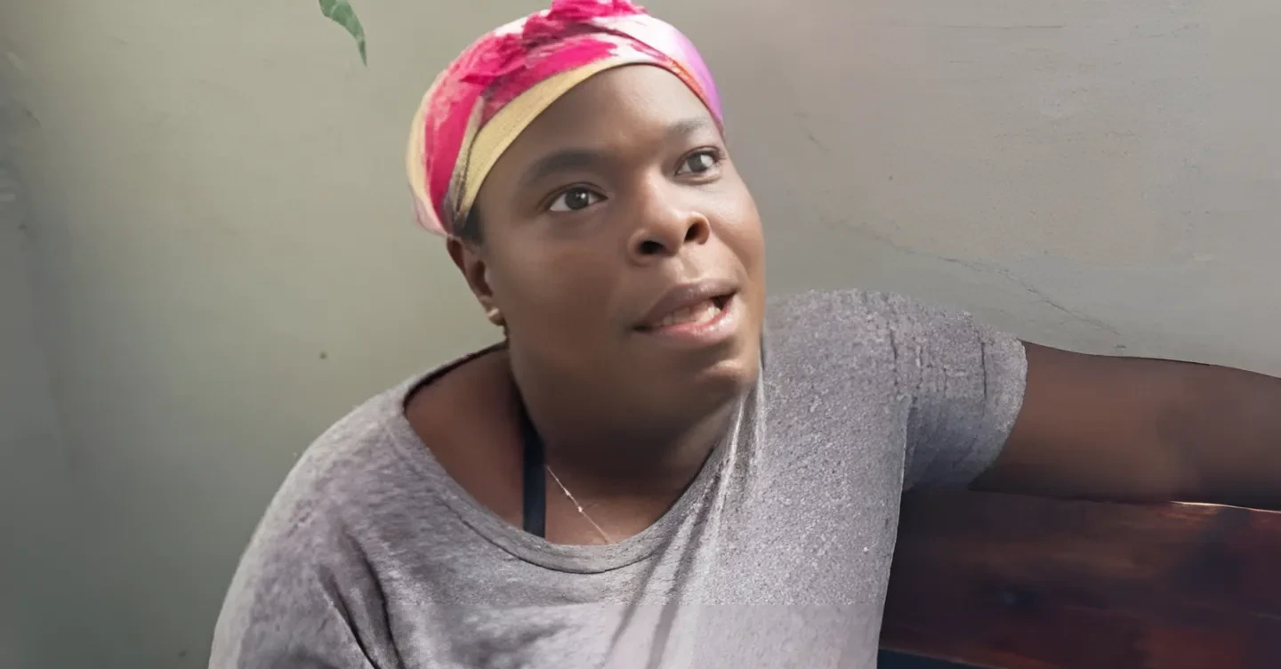 Woman wearing a pink and yellow head wrap sits on a bench, speaking or explaining something to the viewer.