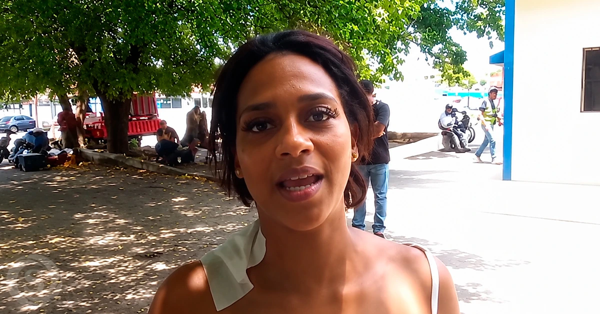 Woman talking to the camera outdoors, with a tree-lined area and people in the background.